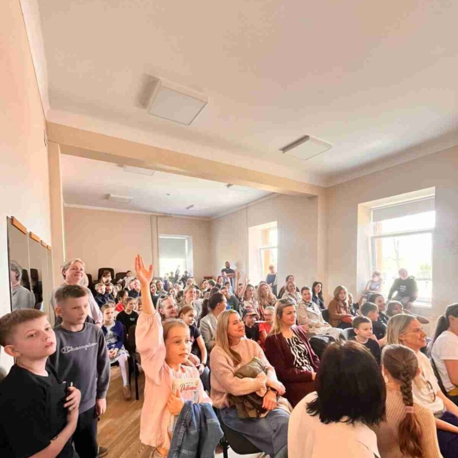 Jauna meitene rozā kreklā stāv gaišā telpā un paceļ roku, kamēr apkārt sēž citi pieaugušie un bērni.