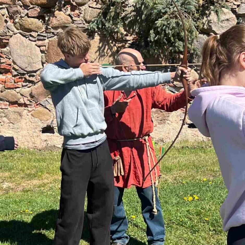 Jauns vīrietis pelēkā džemperī ar kapuci mācās šaut ar loku, kamēr vecāks vīrietis sarkanā tunikā palīdz viņam turēt loku un mērķēt. Fonā redzama akmens siena, un viņi stāv uz zaļas zāles.