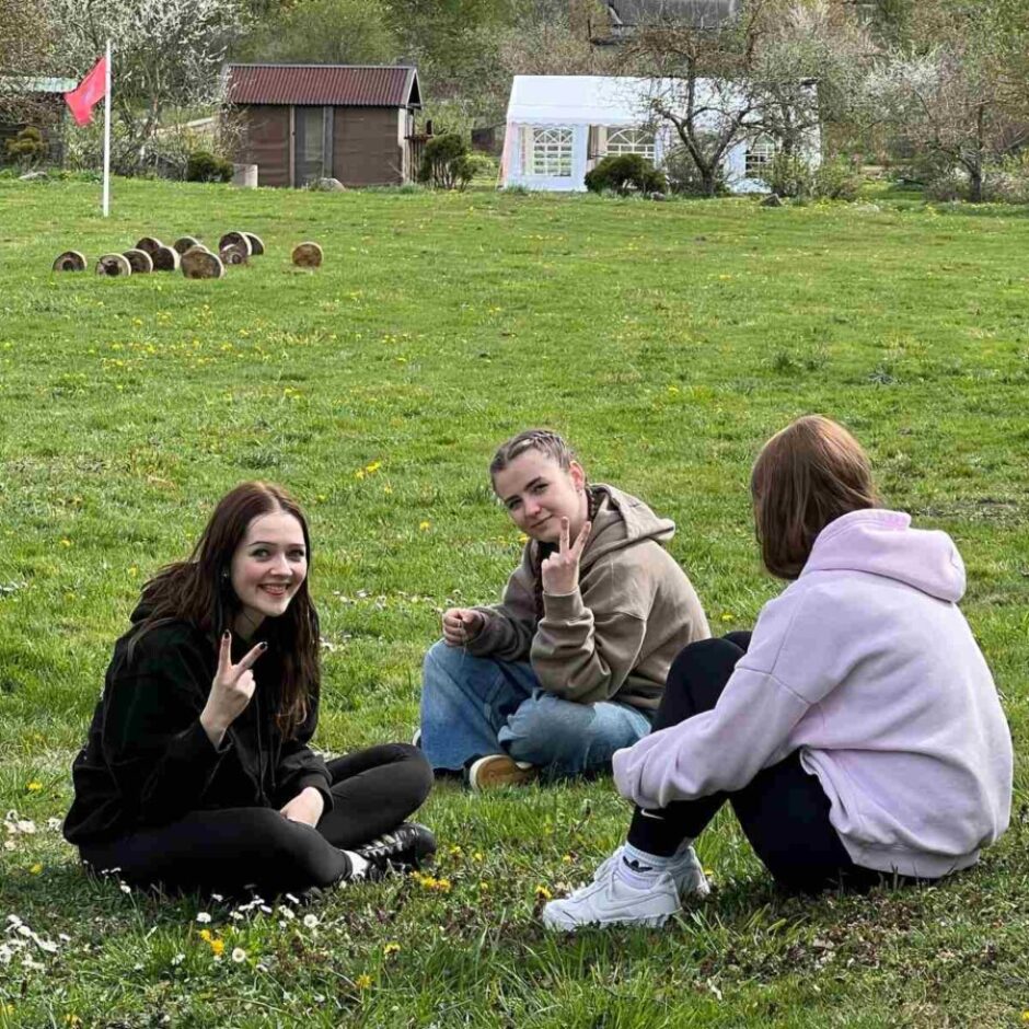 Trīs jaunas sievietes sēž zaļā pļavā ar dzeltenām pienenēm. Divas no viņām skatās kamerā, smaidot un rādot miera zīmi ar pirkstiem. Trešā sieviete redzama no mugurpuses. Fonā redzams sarkans karogs, koka baļķi, neliela koka būda un balta telts.
