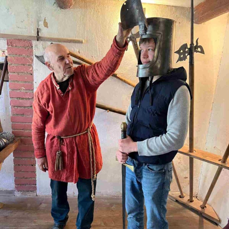 Vīrietis sarkanā tunikā paceļ viduslaiku ķiveres vizieri zēnam, kurš to valkā un tur rokās zobenu. Aizmugurē redzami vēsturiski ieroči un koka grīda.