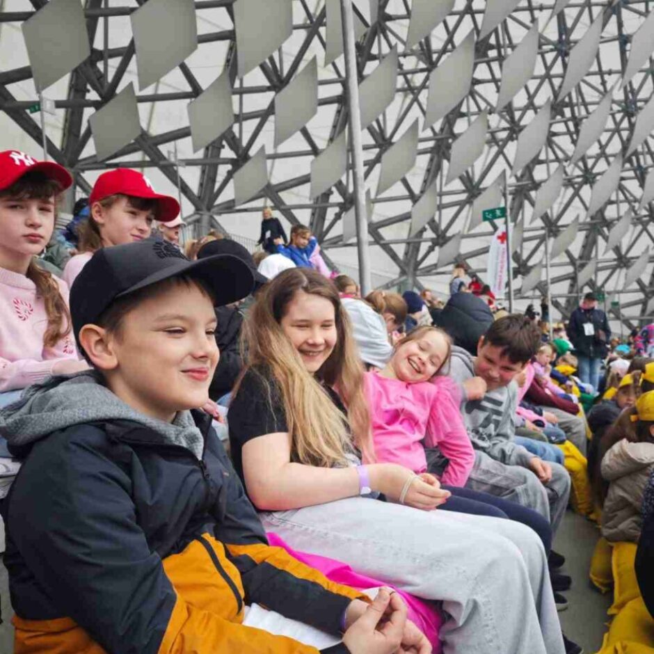 Grupa bērnu sēž stadionam līdzīgās tribīnēs zem moderna, ģeometriska griestu dizaina. Priekšplānā zēns melnā cepurē un dzelteni-melnā jakā viegli smaida, blakus viņam meitene ar gariem blondiem matiem plaši smaida, un cita meitene rozā kreklā smejas, atspiedusies pret viņu. Aizmugurē redzami arī citi priecīgi bērni. Sēdvietas ir dzeltenas, un plašajā norises vietā ir daudz cilvēku.