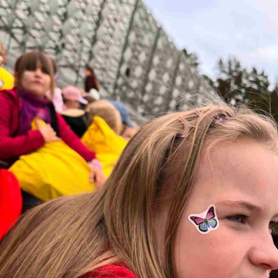 Smaidīga jauna meitene ar gariem blondiem matiem un rozā matu sprādzēm, sarkanā džemperī. Viņai uz vaiga ir zili rozā tauriņa uzlīme. Fonā redzami citi cilvēki dzeltenā apģērbā un metāla konstrukcija.