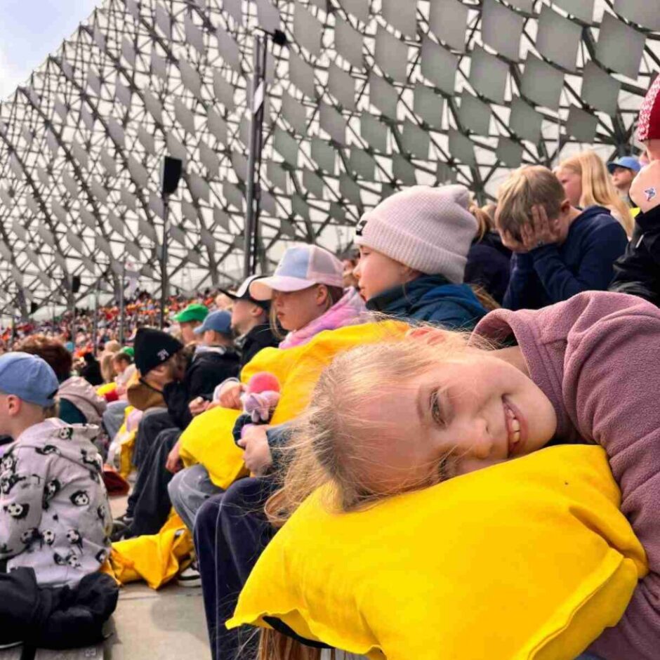 Jauna meitene ar gaišiem matiem smaidot atpūšas, galvu nolikusi uz spilgti dzeltena spilvena. Viņa sēž pūlī blakus citiem bērniem modernā stadiona tribīnēs.