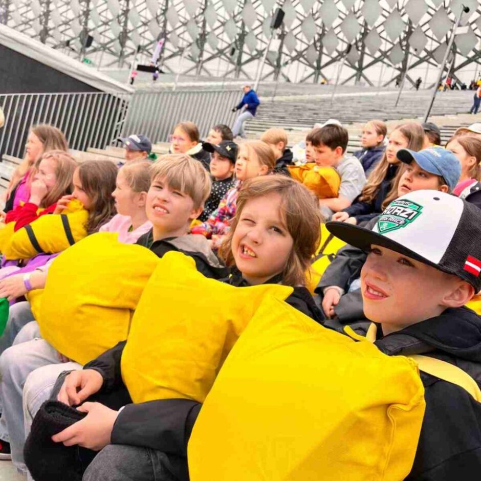 Bērnu grupa sēž moderna stadiona tribīnēs, daudzi tur dzeltenus spilvenus. Priekšplānā redzams zēns cepurē ar uzrakstu 