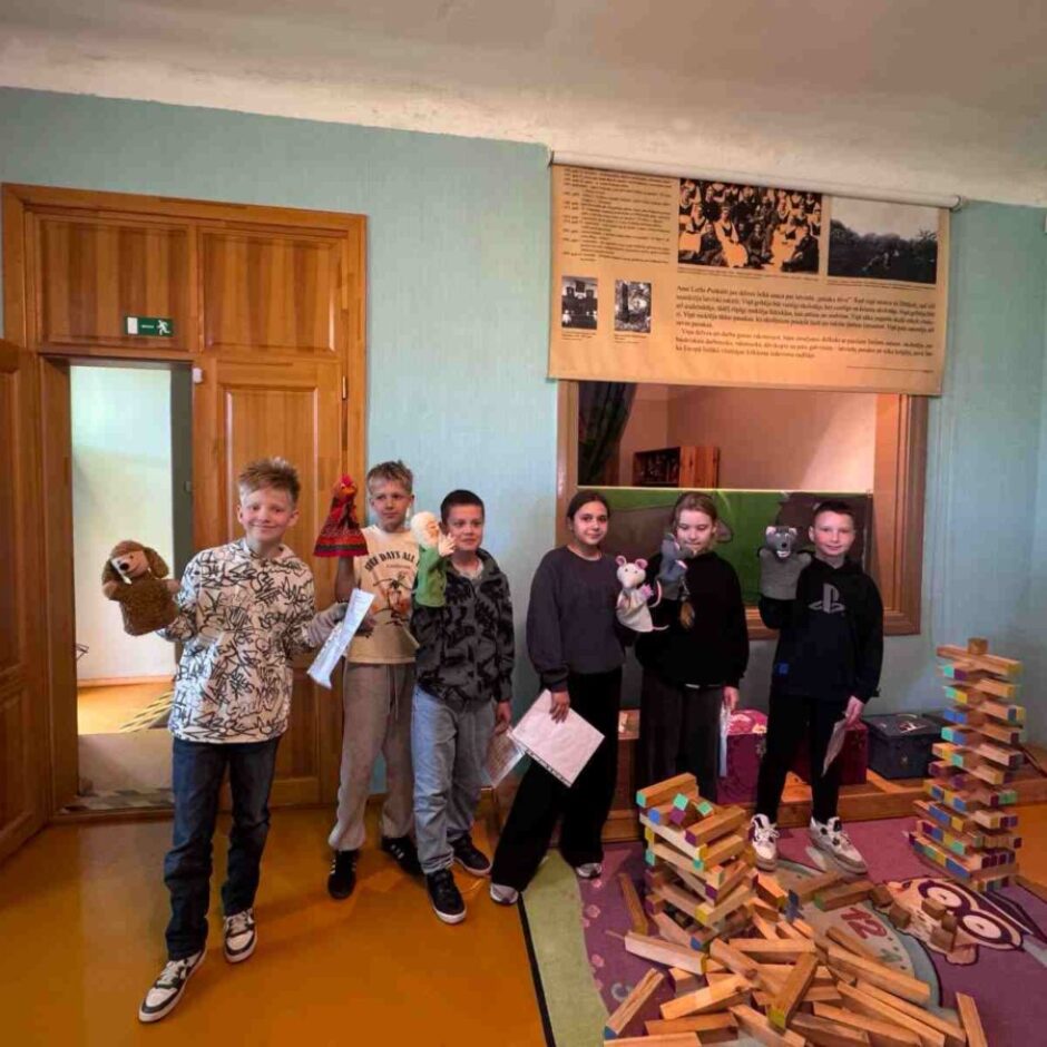Seši bērni, turot rokās lelles, smaidot stāv gaišzilā telpā ar koka durvīm. Viņi tur brūnu suņa lelli, sarkanu figūras lelli, peles un žurkas lelles. Virs padziļināta loga ir liels auduma plakāts ar vēsturiskiem attēliem un tekstu. Uz grīdas ir daudz koka klucīšu, daži no tiem sakrauti torņos.