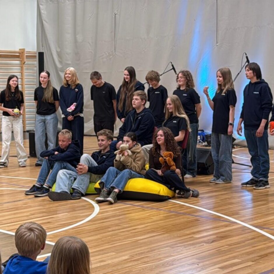 Sporta zālē grupa pusaudžu stāv un sēž uz pupu maisiem, daži turot bumbu vai plīša rotaļlietas, kamēr priekšā sēž jaunāki bērni. Aizmugurē redzams skatuves apgaismojums uz gaiša fona.