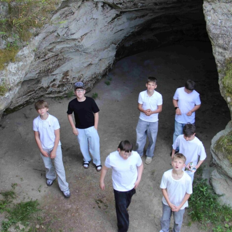 Septiņi zēni stāv pie alas ieejas un skatās augšup. Alas akmeņainās sienas klātas ar sūnām un tajās iegravēts teksts, tostarp vārds 