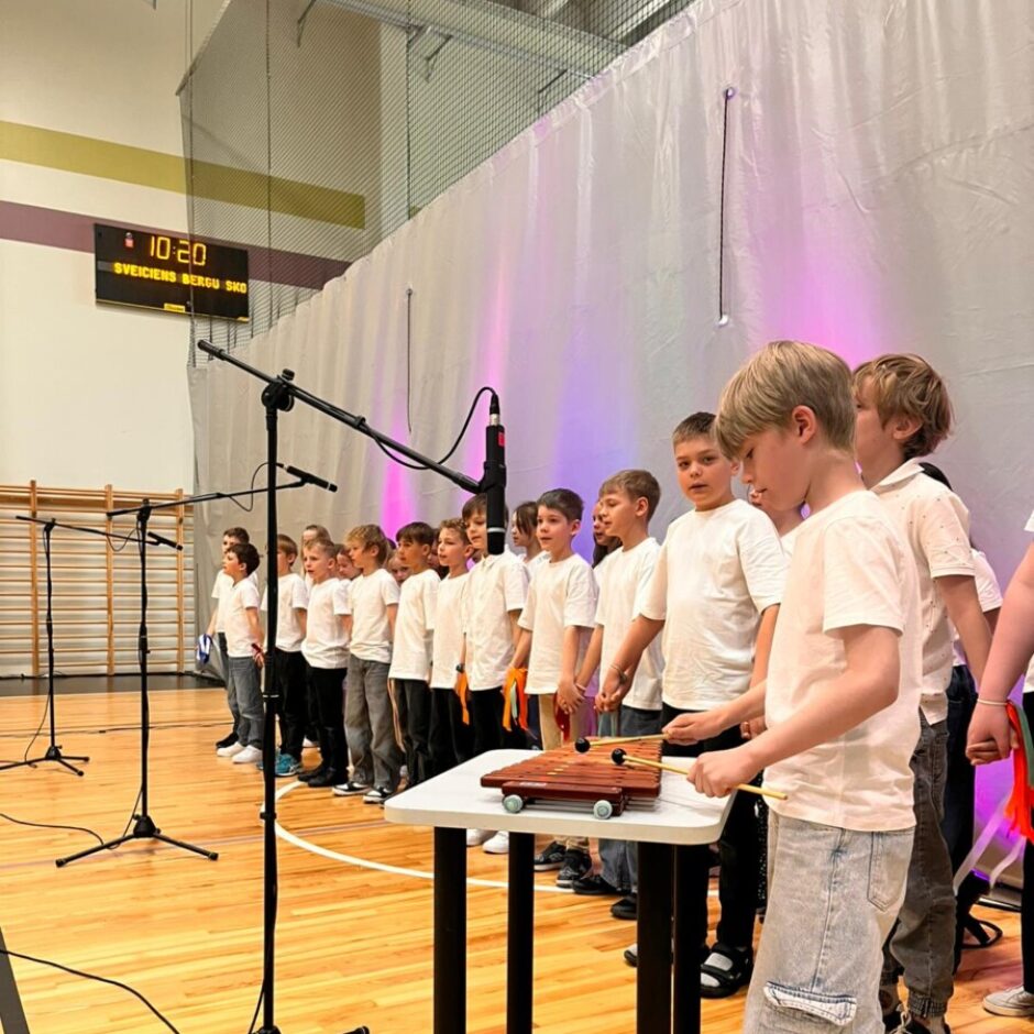 Bērnu grupa sporta zālē uzstājas ar dziesmām un mūziku. Zēni baltos T-kreklos stāv rindā pie mikrofoniem, bet viens zēns priekšplānā spēlē koka ksilofonu. Virs baltā fona, ko apgaismo violeta gaisma, redzams tablo ar uzrakstu SVEICIENS BERGU SKOLA.