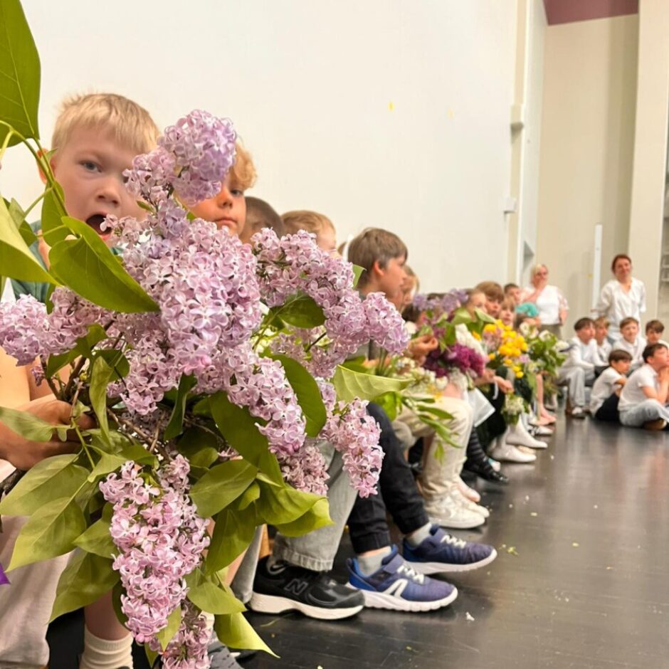 Bērnu rinda sēž uz tumšas skatuves, turot ziedu pušķus. Priekšplānā zēns ar atvērtu muti tur lielu ceriņu pušķi, aiz viņa redzami citi bērni ar dažādu krāsu, tostarp ceriņu, ziediem. Fonā stāv pieaugušie.