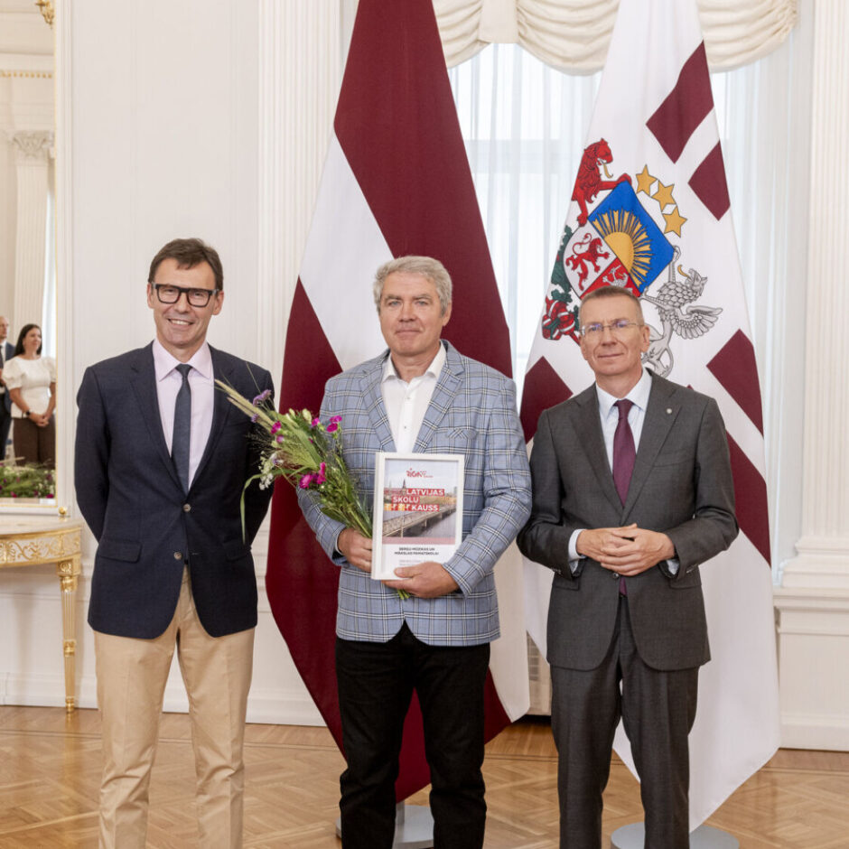 Trīs vīrieši, tostarp sporta skolotājs Oskars un valsts prezidents E.Rinkēvičs, pozē fotogrāfijai formālā telpā ar diviem Latvijas karogiem fonā.