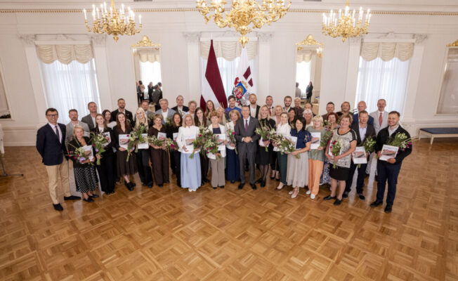 Liela aptuveni piecdesmit cilvēku grupa, kas sastāv no vīriešiem un sievietēm, stāv greznā zālē ar parketa grīdu, turot rokās ziedu pušķus un grāmatas vai sertifikātus. Grupas centrā atrodas vīrietis uzvalkā. Fonā redzams Latvijas valsts karogs un cits valsts simbolikas karogs, bet virs galvas – kristāla lustras.