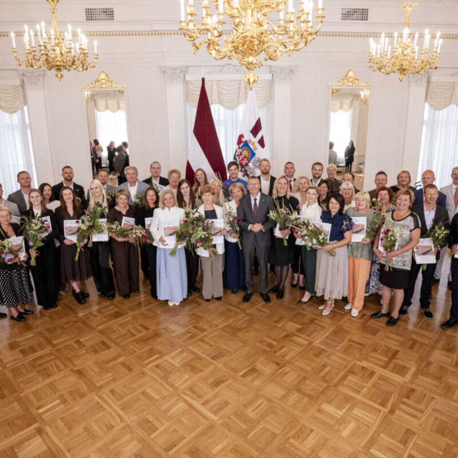 Liela aptuveni piecdesmit cilvēku grupa, kas sastāv no vīriešiem un sievietēm, stāv greznā zālē ar parketa grīdu, turot rokās ziedu pušķus un grāmatas vai sertifikātus. Grupas centrā atrodas vīrietis uzvalkā. Fonā redzams Latvijas valsts karogs un cits valsts simbolikas karogs, bet virs galvas – kristāla lustras.