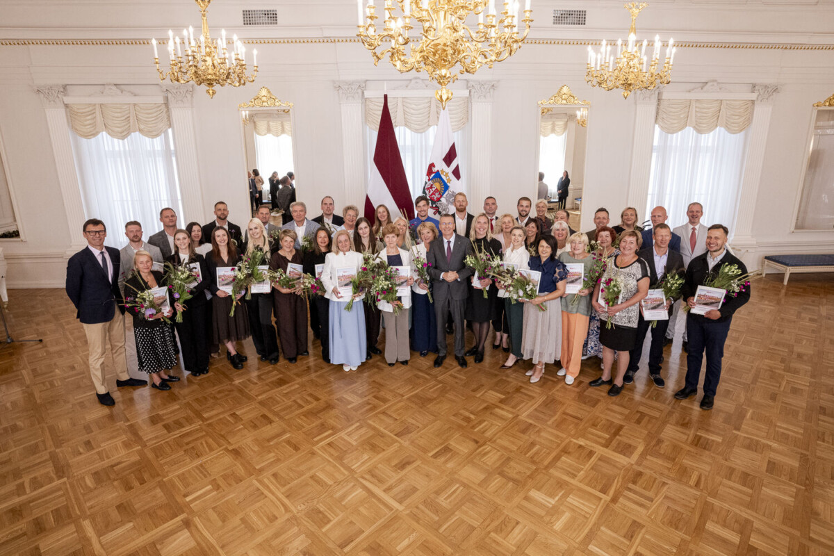 Liela aptuveni piecdesmit cilvēku grupa, kas sastāv no vīriešiem un sievietēm, stāv greznā zālē ar parketa grīdu, turot rokās ziedu pušķus un grāmatas vai sertifikātus. Grupas centrā atrodas vīrietis uzvalkā. Fonā redzams Latvijas valsts karogs un cits valsts simbolikas karogs, bet virs galvas – kristāla lustras.
