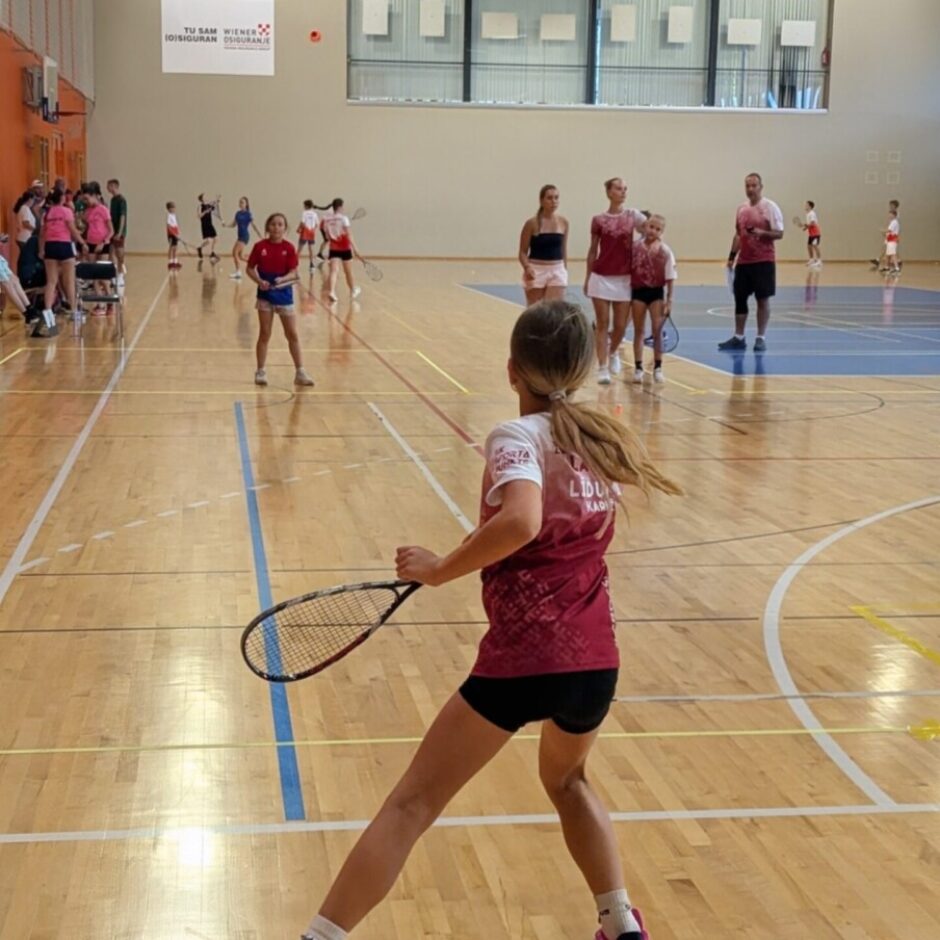 Jauna meitene no muguras, sporta zālē ar raketi rokā. Viņa valkā bordo kreklu, melnus šortus un rozā sporta apavus. Fonā redzami citi bērni un pieaugušie ar raketēm.