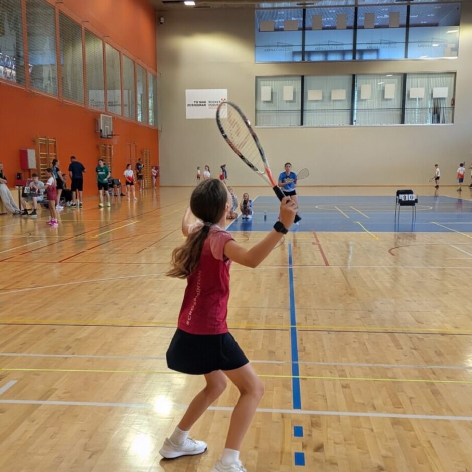 Jauna meitene sarkanā kreklā un melnā svārkos, ar krosmintona raketi rokā, gatavojas sacensībai sporta zālē. Viņa stāv uz koka grīdas ar laukuma līnijām, fonā redzami citi spēlētāji, tablo ar 
