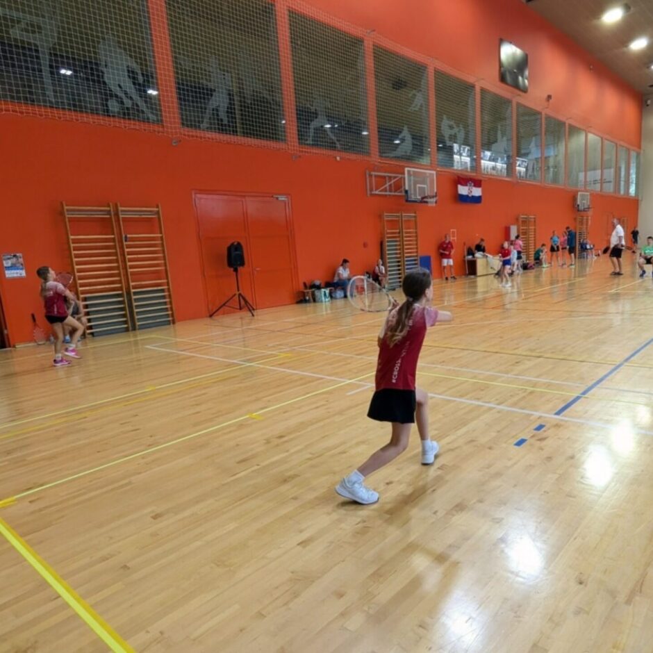 Jauna meitene sarkanā kreklā un melnos svārkos gatavojas spēlēt badmintonu koka laukuma priekšplānā. Iekštelpu sporta zālē ar oranžām sienām fonā redzami vairāki citi cilvēki, kas spēlē krosmintonu.