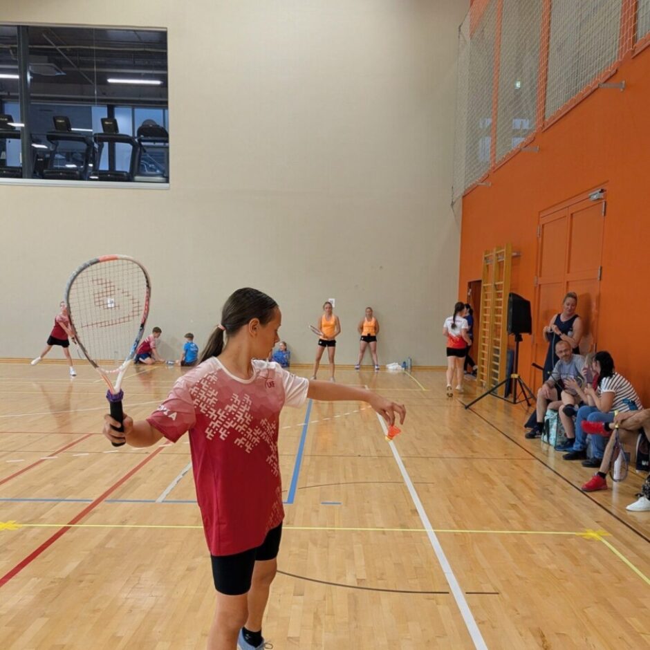 Jauna meitene sporta zālē tur rokās krosminton raketi un oranžu volānu, gatavojoties servēt. Viņa valkā sarkanu kreklu ar baltiem rakstiem un 