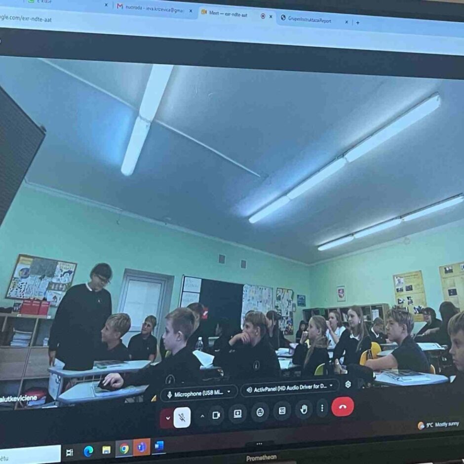 Datora ekrānā redzams Google Meet videozvans, kurā attēlota skolas klase ar skolēniem un skolotāju, kas mijiedarbojas mācību stundas laikā. Videozvana iekšpusē, apakšējā labajā stūrī, ir mazāks attēls ar to pašu klasi. Kā dalībnieks norādīta 