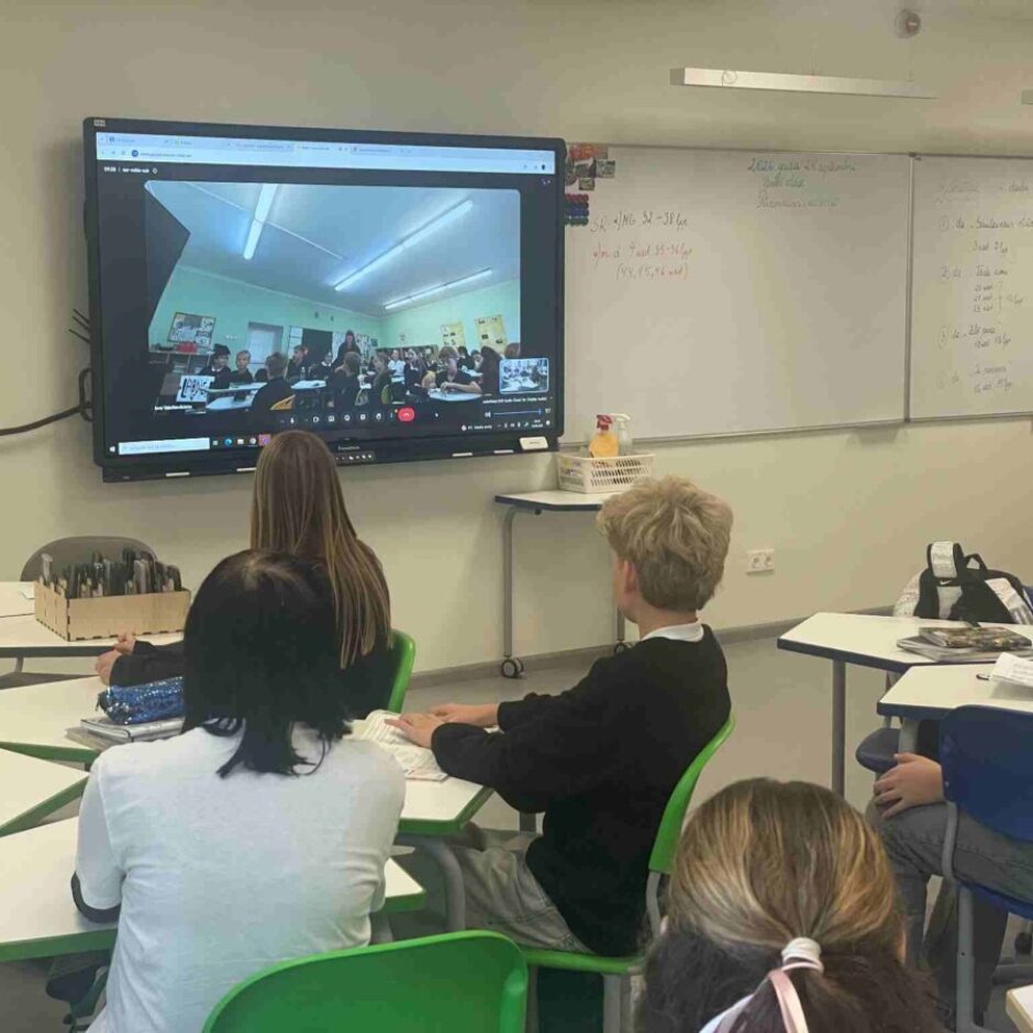 Klasē skolēni sēž pie galdiem un skatās uz lielu interaktīvo ekrānu, kurā redzama videokonference ar citu klasi, kas arī pilna ar skolēniem.