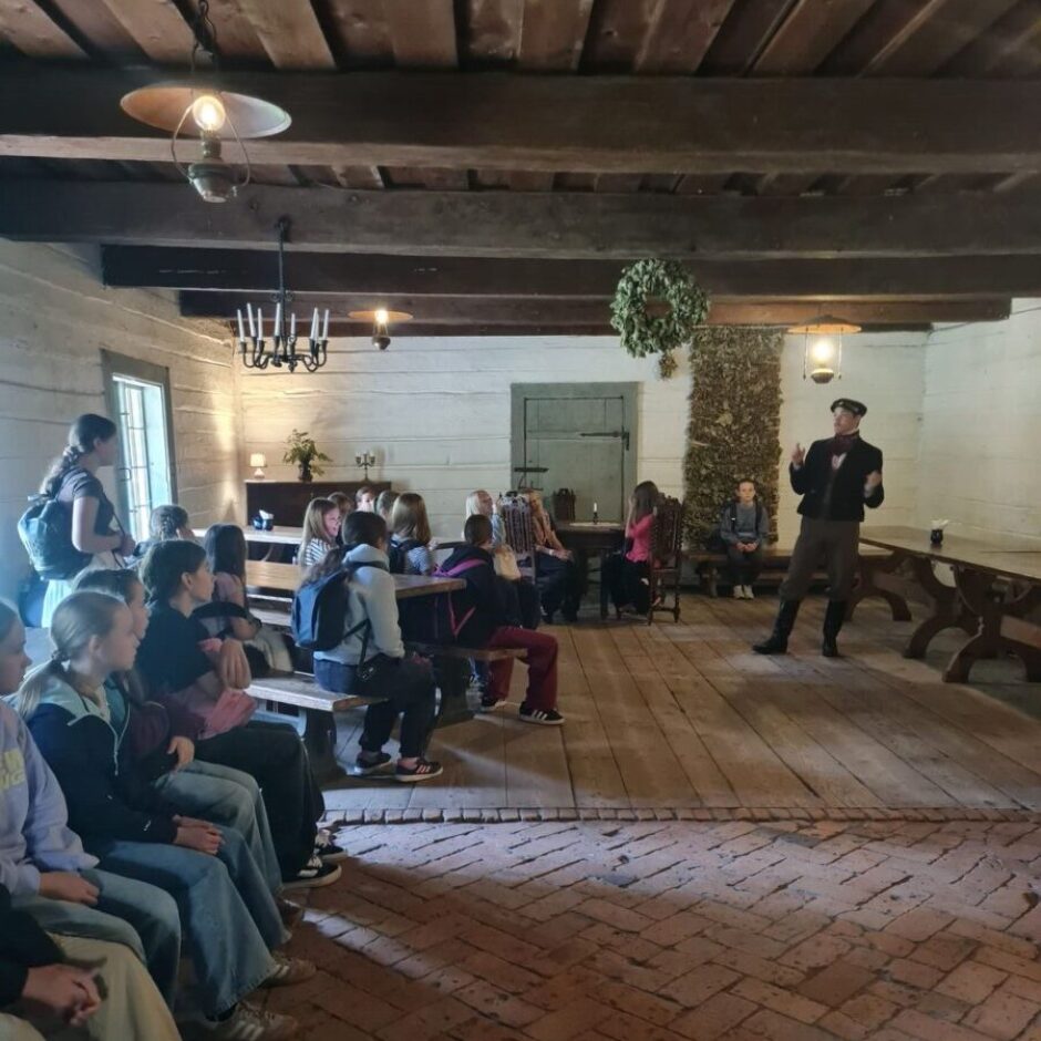 Vēsturiskā koka telpā ar ķieģeļu grīdu vīrietis vēsturiskā tērpā stāv un runā, žestikulējot ar rokām, savukārt skolēnu grupa sēž un klausās viņu.