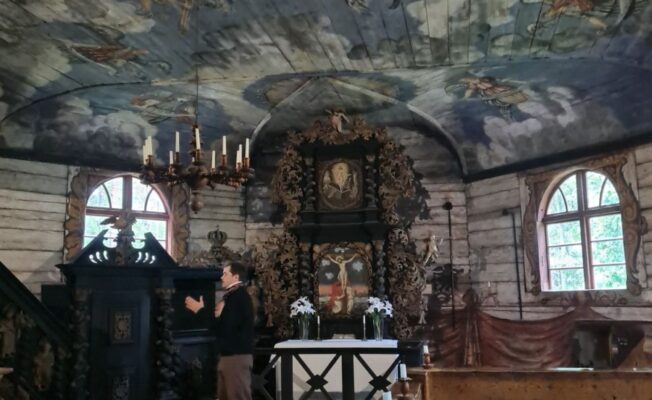 Vecas koka baznīcas interjers ar bagātīgi apgleznotiem griestiem, kuros redzami mākoņi un debesu figūras. Vēsturiskā tērpā ģērbts manekens stāv uz sarkana paklāja pie grezna tumša koka altāra, uz kura ir krucifikss un glezna. Priekšplānā koka solā sēž jauna sieviete.
