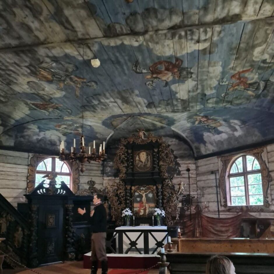 Vecas koka baznīcas interjers ar bagātīgi apgleznotiem griestiem, kuros redzami mākoņi un debesu figūras. Vēsturiskā tērpā ģērbts manekens stāv uz sarkana paklāja pie grezna tumša koka altāra, uz kura ir krucifikss un glezna. Priekšplānā koka solā sēž jauna sieviete.