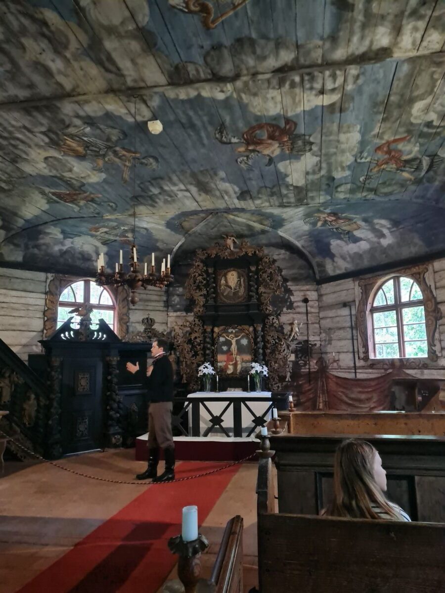 Vecas koka baznīcas interjers ar bagātīgi apgleznotiem griestiem, kuros redzami mākoņi un debesu figūras. Vēsturiskā tērpā ģērbts manekens stāv uz sarkana paklāja pie grezna tumša koka altāra, uz kura ir krucifikss un glezna. Priekšplānā koka solā sēž jauna sieviete.