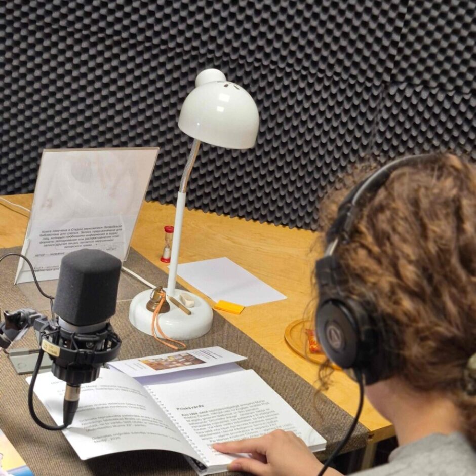 Cilvēks ar austiņām sēž ierakstu studijā pie mikrofona, lasot atvērtu grāmatu, kuras lapā redzams virsraksts 