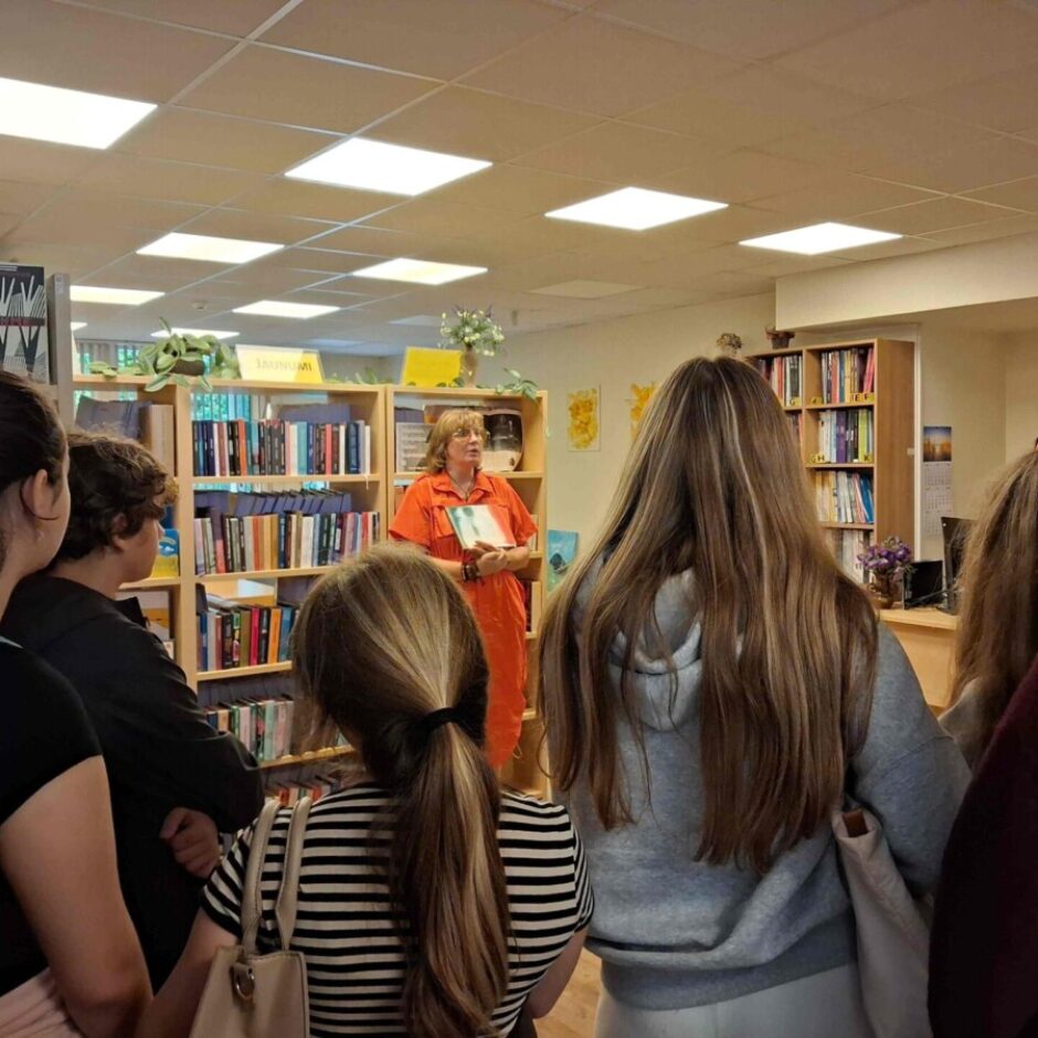 Sieviete oranžā tērpā runā ar jauniešu grupu bibliotēkā vai grāmatnīcā, turot rokās grāmatu. Jaunieši stāv viņai priekšā ar mugurām pret skatītāju un klausās. Fonā redzami grāmatu plaukti, un uz dzeltenas zīmes lasāms teksts 
