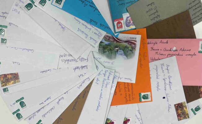 Vairākas krāsainas aploksnes, izklātas vēdekļveidā uz galda. Aploksnes ir baltas, zilas, oranžas, brūnas un citas, un uz tām redzamas Latvijas pastmarkas. Uz dažām aploksnēm ir rakstītas adreses