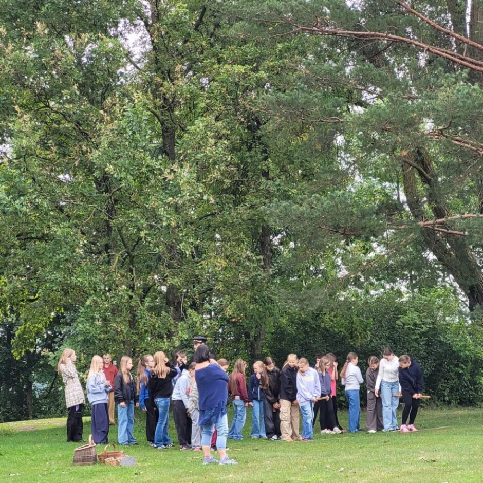 Jauniešu grupa stāv rindā zaļā pļavā ar blīvu koku fonu. Sieviete zilā kreklā stāv pretī grupai, vadot āra aktivitāti, kurā uz zemes izkaisīti koka zari.