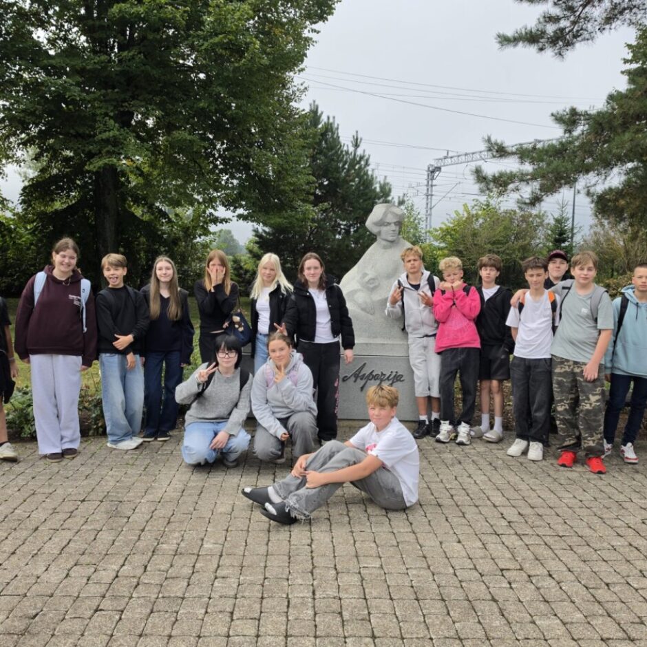 Aptuveni 18 pusaudži un bērni pozē āra fotoattēlā pie pieminekļa. Lielākā daļa stāv, daži pietupušies vai sēž uz zemes. Piemineklis ir sievietes krūšutēls ar uzrakstu 