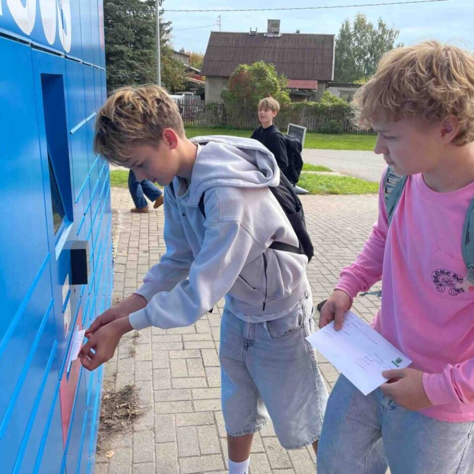Divi jaunieši stāv pie zila automāta. Viens ievieto aploksni atverē, bet otrs tur vēl vienu aploksni. Uz automāta redzams daļējs teksts: 