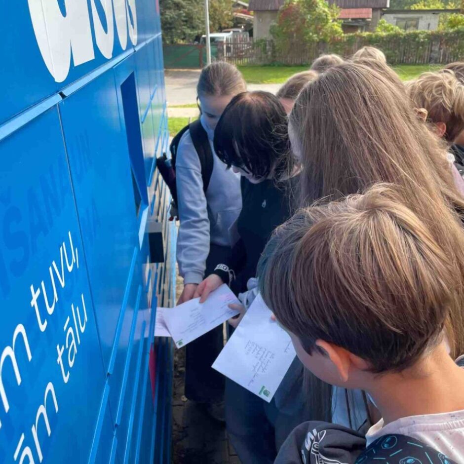 Skolēnu grupa pulcējusies ap zilu konteineru vai automātu, uz kura redzams uzraksts 