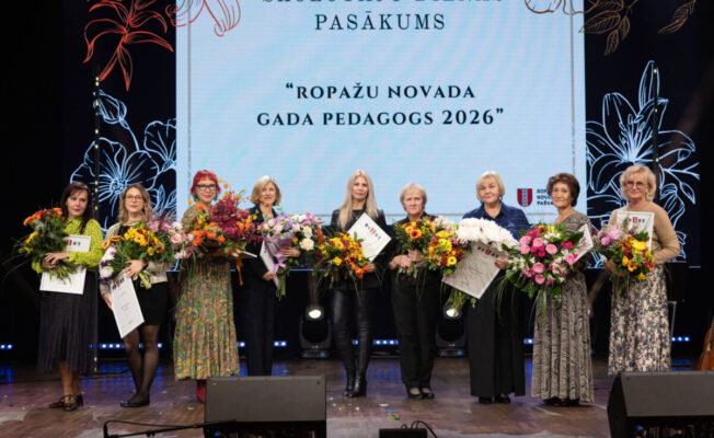 Deviņas sievietes stāv uz skatuves, turot krāšņus ziedu pušķus un sertifikātus. Aiz viņām redzams liels ekrāns ar uzrakstiem 