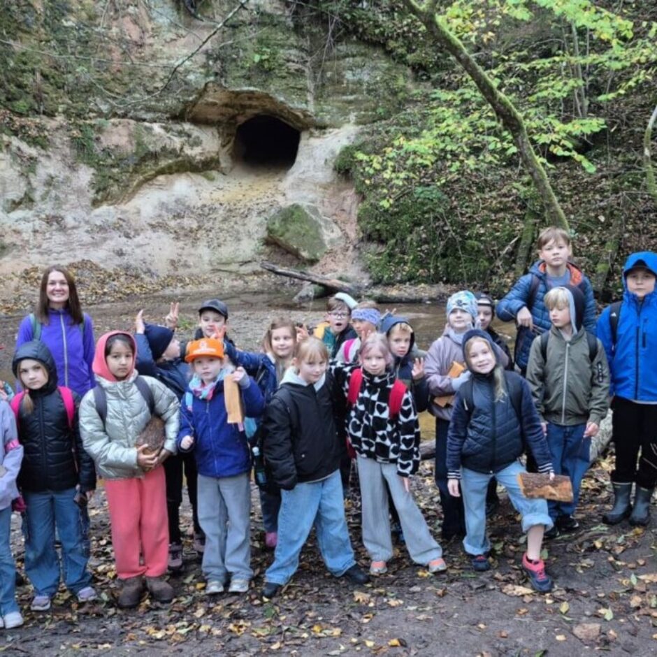 Bērnu grupa ar pieaugušo sievieti stāv pie smilšakmens klints ar alas ieeju pārgājienā dabā. Zeme ir klāta ar nobirušām lapām, un apkārt redzami koki. Pa labi stāv informācijas stends.
