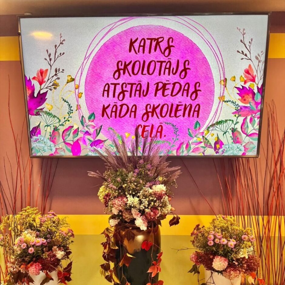 Liels ekrāns rāda uzrakstu rozā aplī ar ziedu motīviem: KATRS SKOLOTĀJS ATSTĀJ PĒDAS KĀDA SKOLĒNA CEĻĀ. Zem ekrāna atrodas trīs lieli puķu podi ar bagātīgiem rudens ziedu un lapu dekoriem; centrā ir tumšzaļš ar zelta akcentiem, sānos divi balti, visi rotāti ar sarkanīgām rudens lapām.