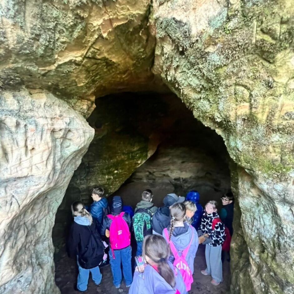 Bērnu grupa ar mugursomām pēta alu ar akmeņainām, ar sūnu klātām sienām un tumšu ieeju tālākā daļā.