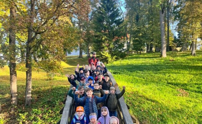 Liela bērnu grupa un pieaugušais stāv uz koka tiltiņa, smaidot un mājot kamerai saulainā rudens dienā. Apkārt tiltiņam ir koki ar dzeltenām un oranžām lapām un zaļa zāle.