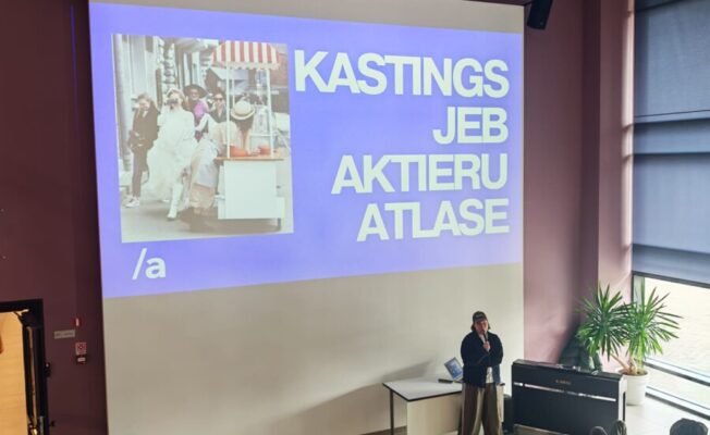 Lektors stāv pie galda ar mikrofonu, prezentējot lielā auditorijā, kur jaunieši sēž uz koka pakāpieniem. Uz liela ekrāna redzams attēls ar cilvēkiem ielā un teksts 