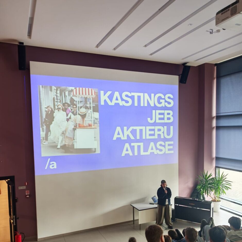 Lektors stāv pie galda ar mikrofonu, prezentējot lielā auditorijā, kur jaunieši sēž uz koka pakāpieniem. Uz liela ekrāna redzams attēls ar cilvēkiem ielā un teksts 