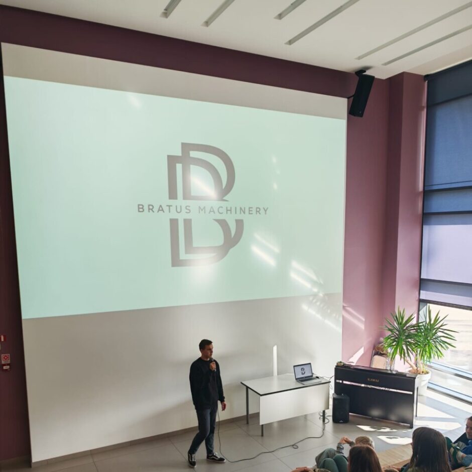 Vīrietis ar mikrofonu stāv auditorijā liela ekrāna priekšā, uz kura redzams logotips un teksts 