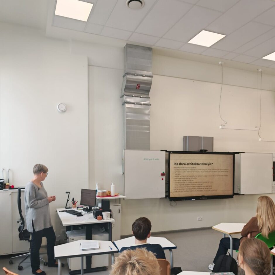 Klasē sieviete stāv pie galda ar datoru, bet vairāki skolēni sēž pie galdiem, vērsti pret lielo ekrānu. Uz ekrāna redzams prezentācijas slaids ar virsrakstu: 
