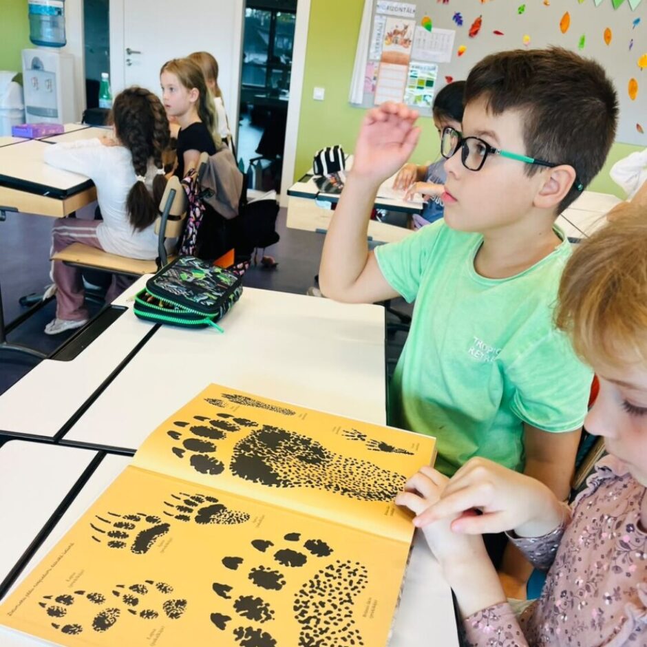 Divi bērni klasē pie galda. Meitene rāda ar pirkstu uz atvērtu grāmatu, kurā attēloti dzīvnieku pēdu nospiedumi ar nosaukumiem lapsa, lūsis, alnis, vilks. Zēns brillēs skatās uz priekšu.