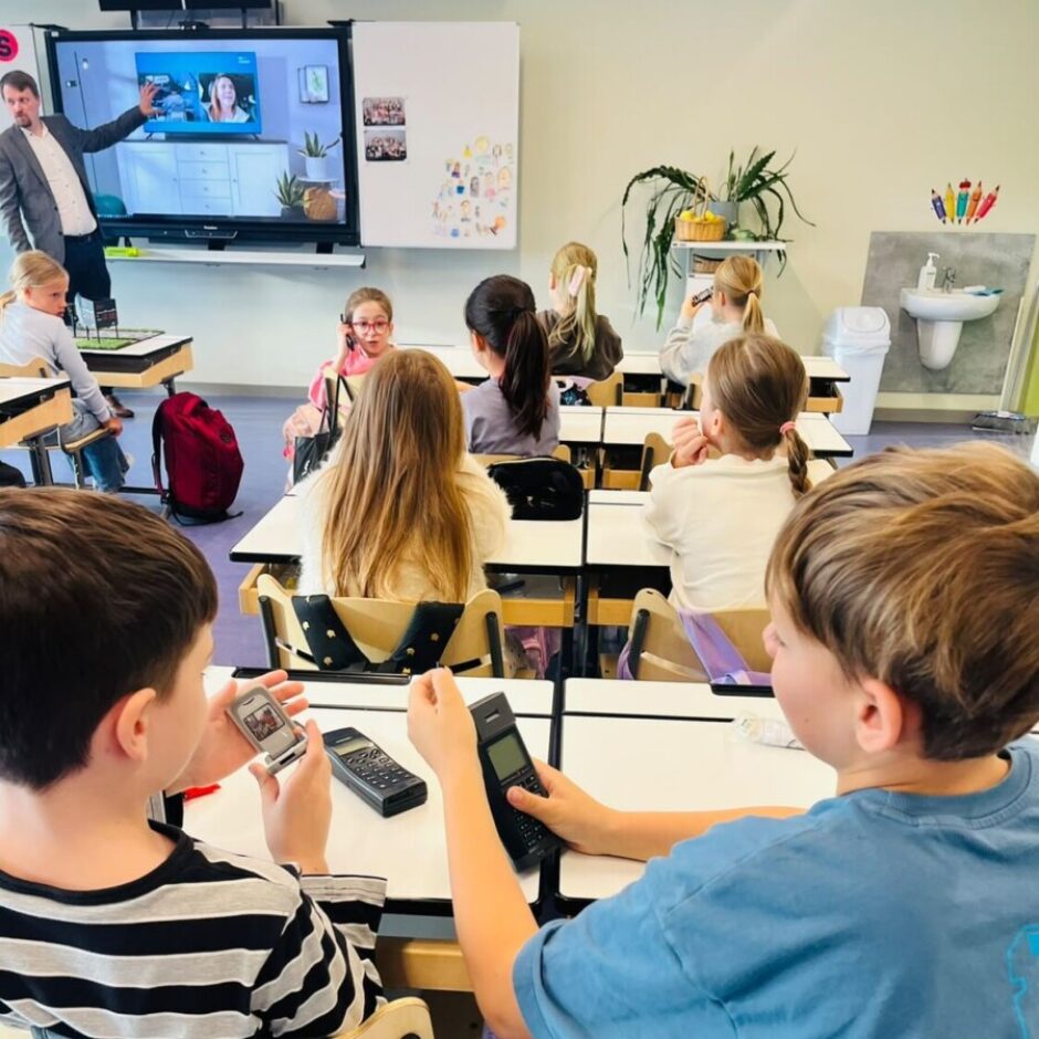 Skolēnu klase, kurā skolotājs rāda uz lielu ekrānu, kas rāda sievieti televizora raidījumā. Priekšplānā divi zēni sēž pie galdiem, turot rokās un pētot vecus mobilos telefonus, iespējams, mācību stundā par tehnoloģijām.