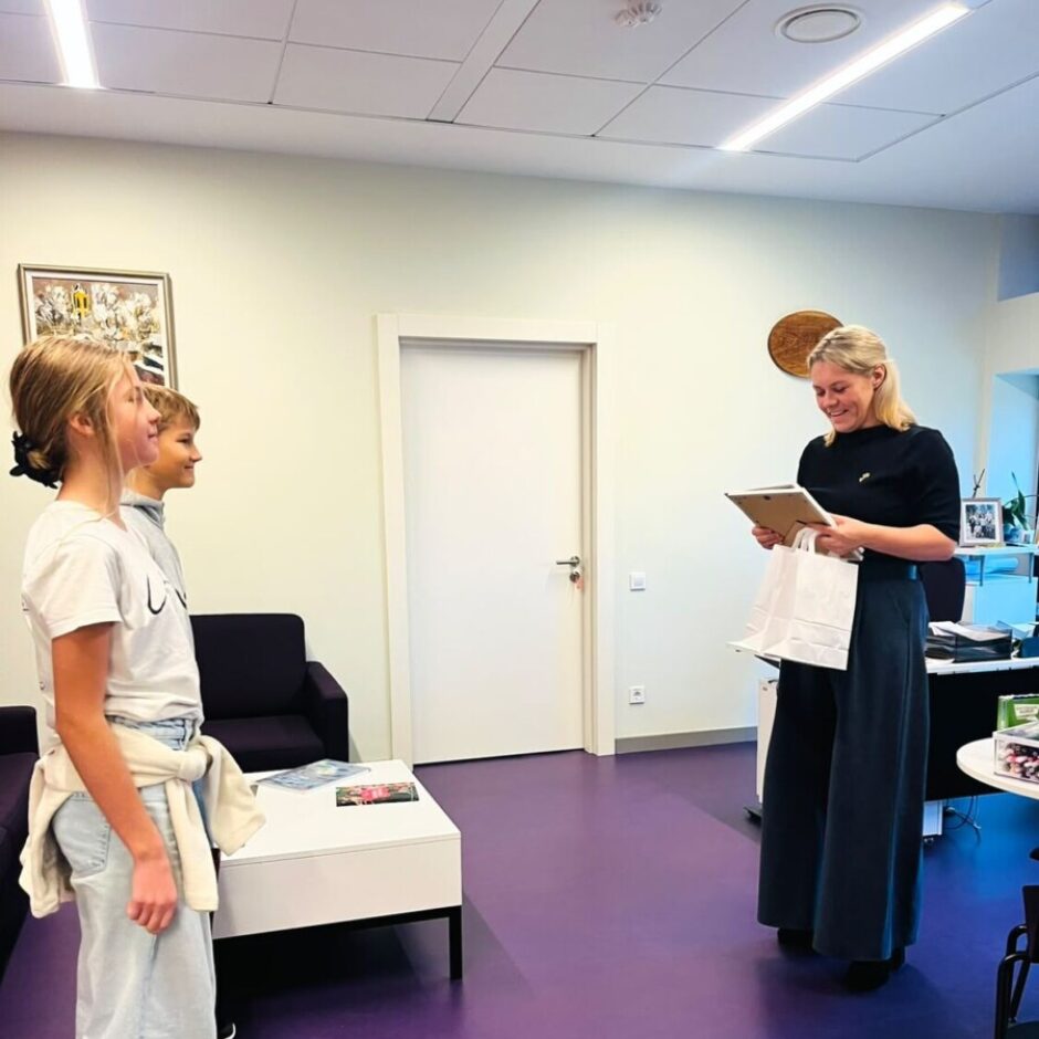 Smaidoša sieviete melnā apģērbā tur rokās ierāmētu priekšmetu un baltu dāvanu maisiņu, stāvot pretī meitenei un zēnam. Viņi atrodas telpā ar violetu grīdu un baltu kafijas galdiņu.