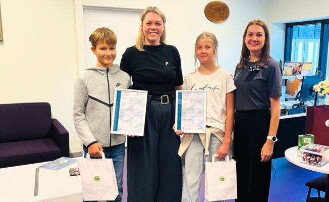 Divas smaidīgas pieaugušas sievietes un divi smaidīgi bērni (zēns un meitene) stāv telpā. Bērni un viena sieviete tur sertifikātus, bet bērni arī mazus baltus dāvanu maisiņus.